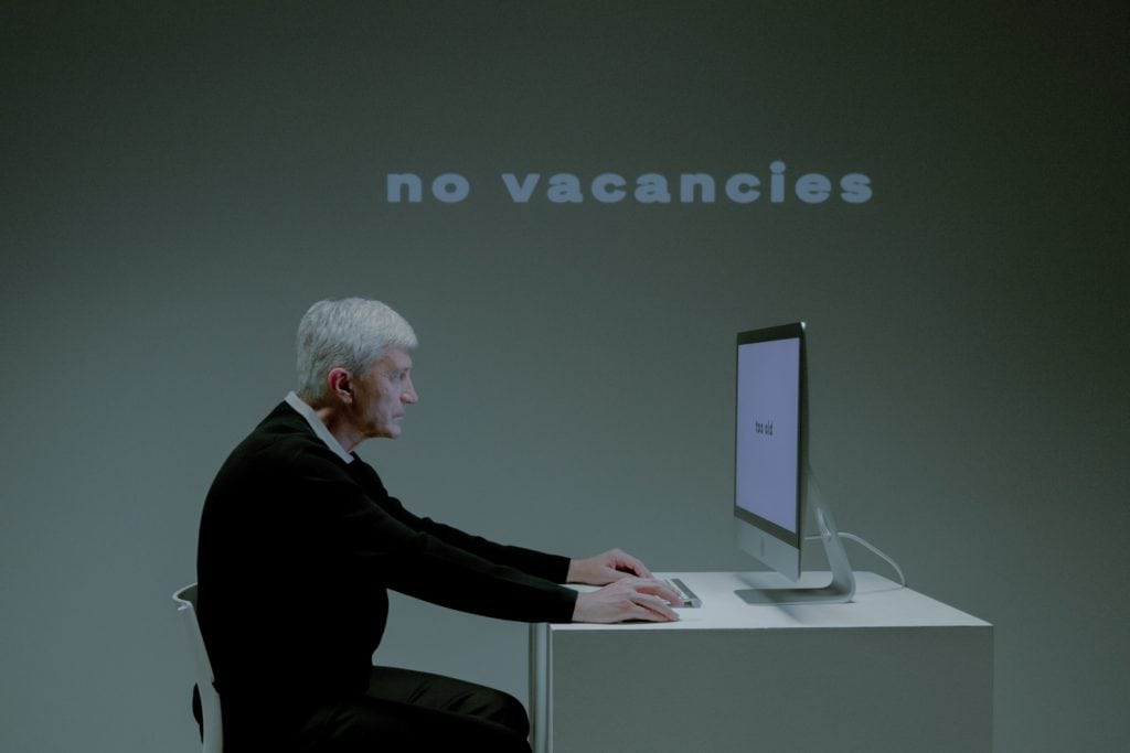 An older person sitting at a desk looking at a computer while the words  "no vacancies" are shown on the wall behind them. Possibly displaying ethical issues similar to not hiring because of older age.