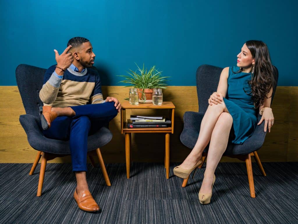 Two people sitting in blue chairs while talking to each other. One has hand gestures indicating they could be engaging in storytelling.