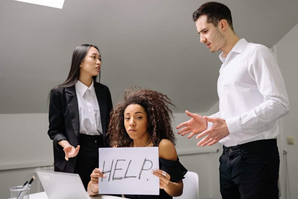 Teo people yelling at another person. The person being yelled at holds up a sign saying "help". An example of conflict in the workplace.