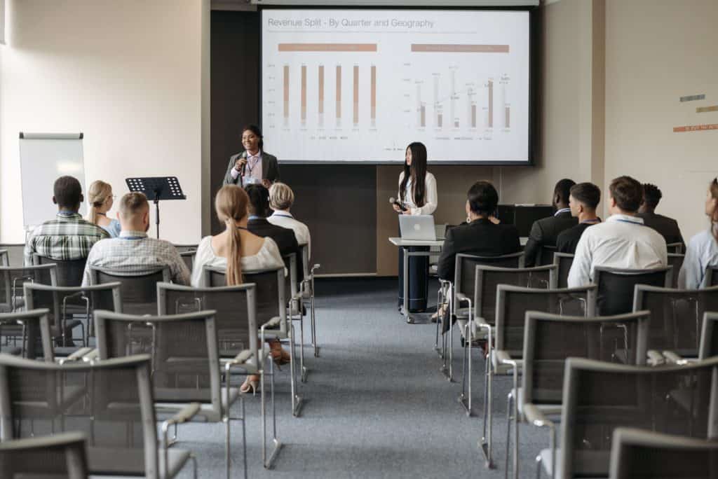 Two people standing at the front of a room, presenting a PowerPoint to others